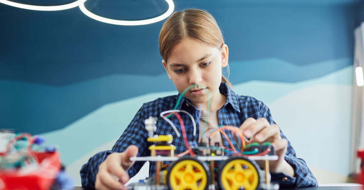 Teenage girl working on a robotic car project with wires and wheels inside a classroom.