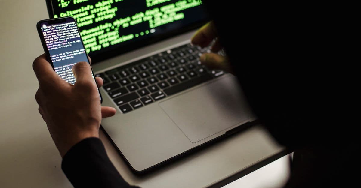 From behind crop male programmer in black hood browsing netbook and mobile phone while working in office