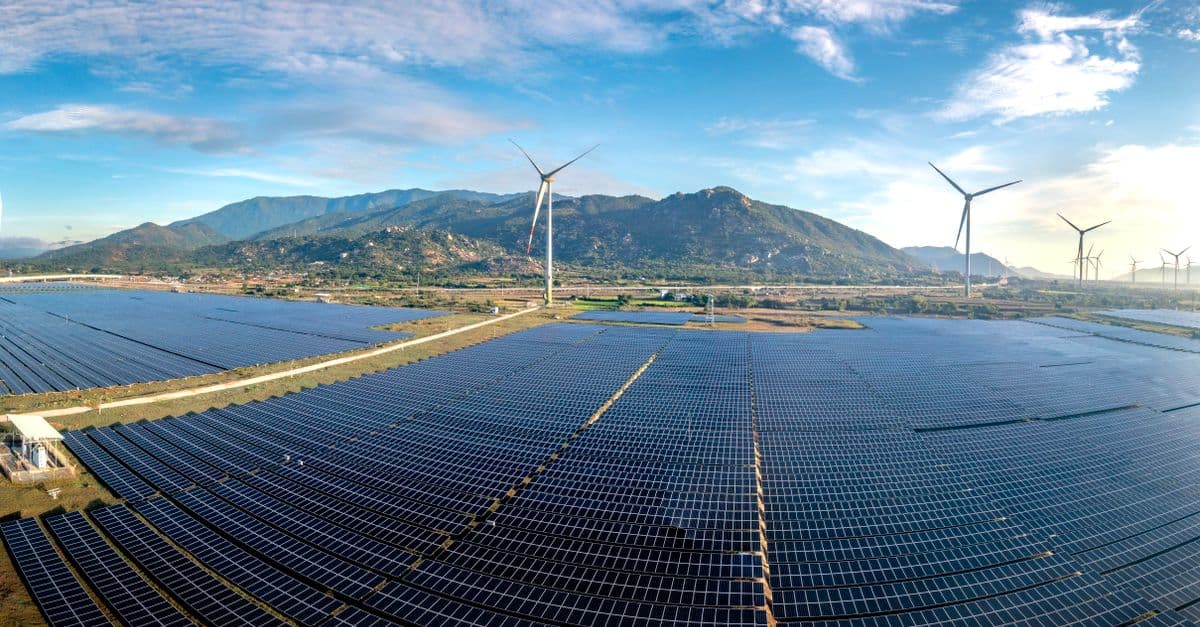 Aerial view of a vast solar farm with wind turbines against a mountainous backdrop on a sunny day.