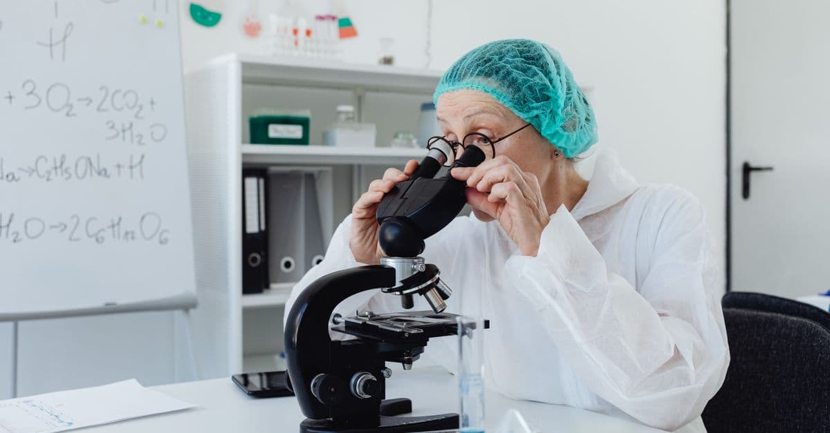 A senior female scientist using a microscope in a laboratory setting, wearing protective gear.