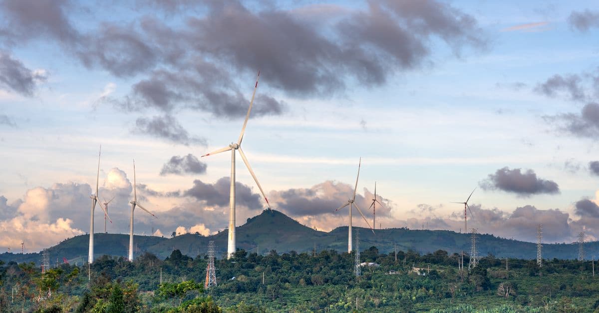 Panoramic view of wind turbines amidst lush greenery and mountains under a cloudy sky.