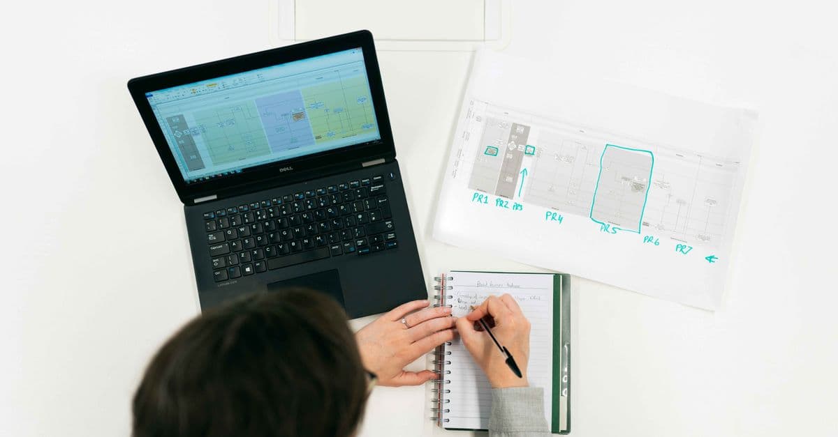 A person working on a laptop, making notes from digital data and charts in an office setting.