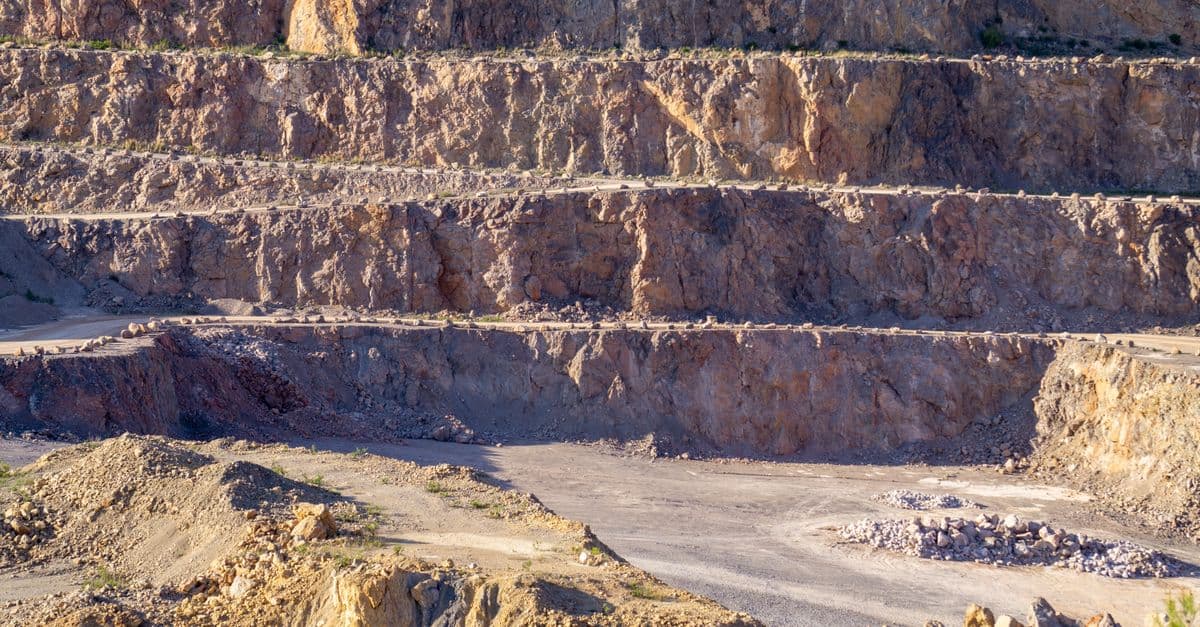 Sunlit view of a rugged, multi-layered rock quarry with exposed strata and rocky terrain.