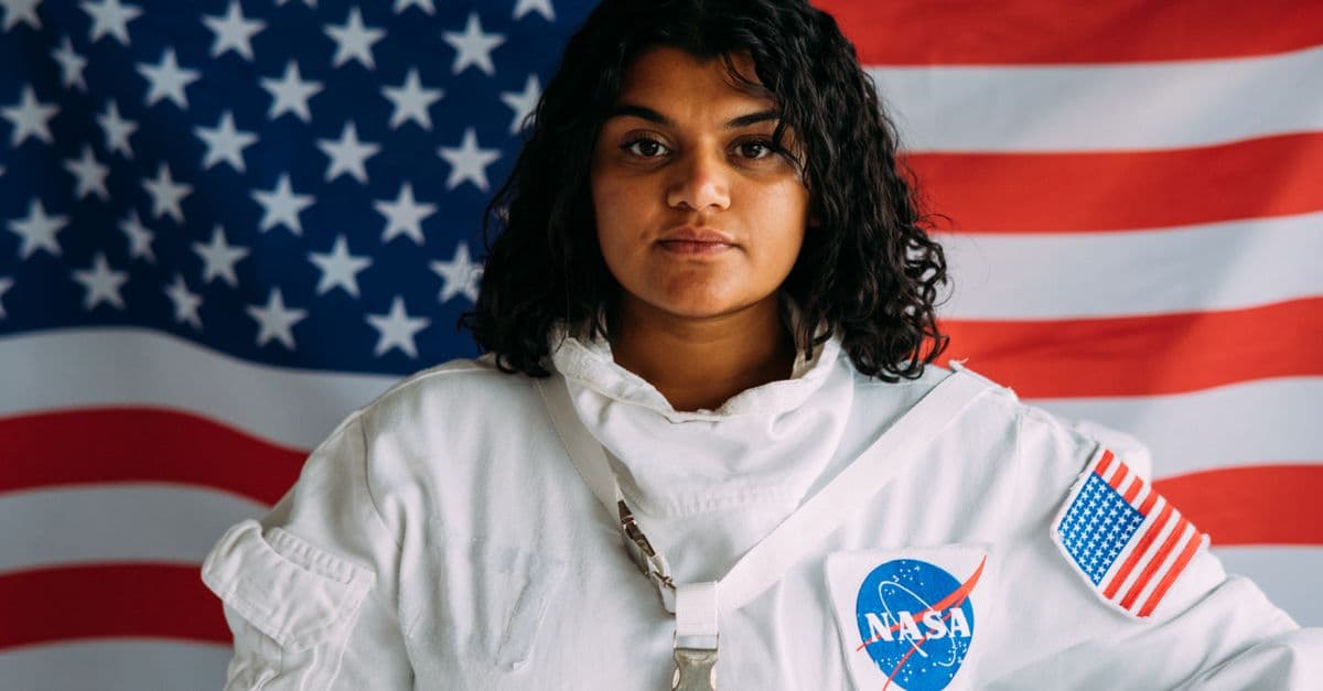A woman in a NASA astronaut suit poses confidently against an American flag background.
