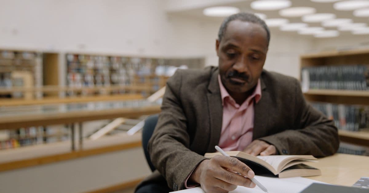 An adult man engaged in reading and writing at a library table, with shelves in the background.