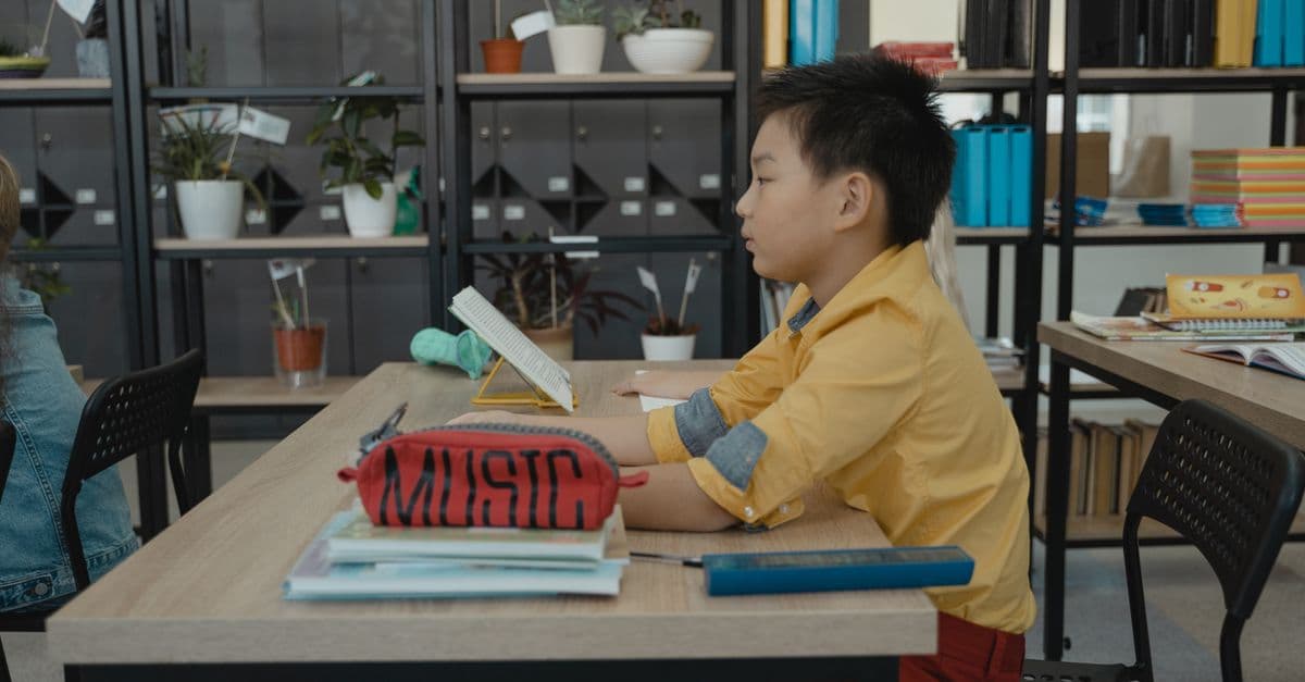 Boy in yellow shirt reading in a classroom with books and plants around.