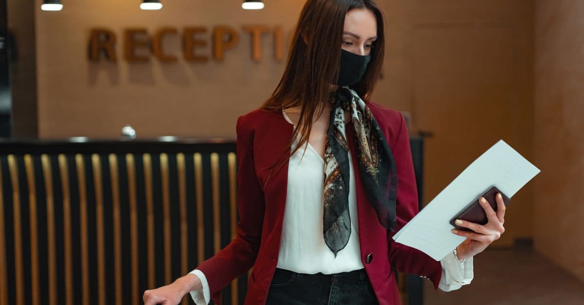 Woman in red blazer at hotel reception, holding luggage and documents, wearing a face mask.