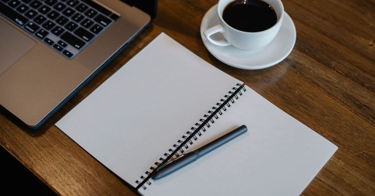 From above of workspace with cup of hot coffee and pen on empty notepad near modern laptop on wooden table