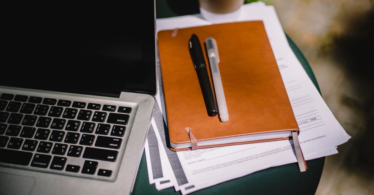 From above of leather notebook with papers and pens placed on table near laptop and cup of coffee