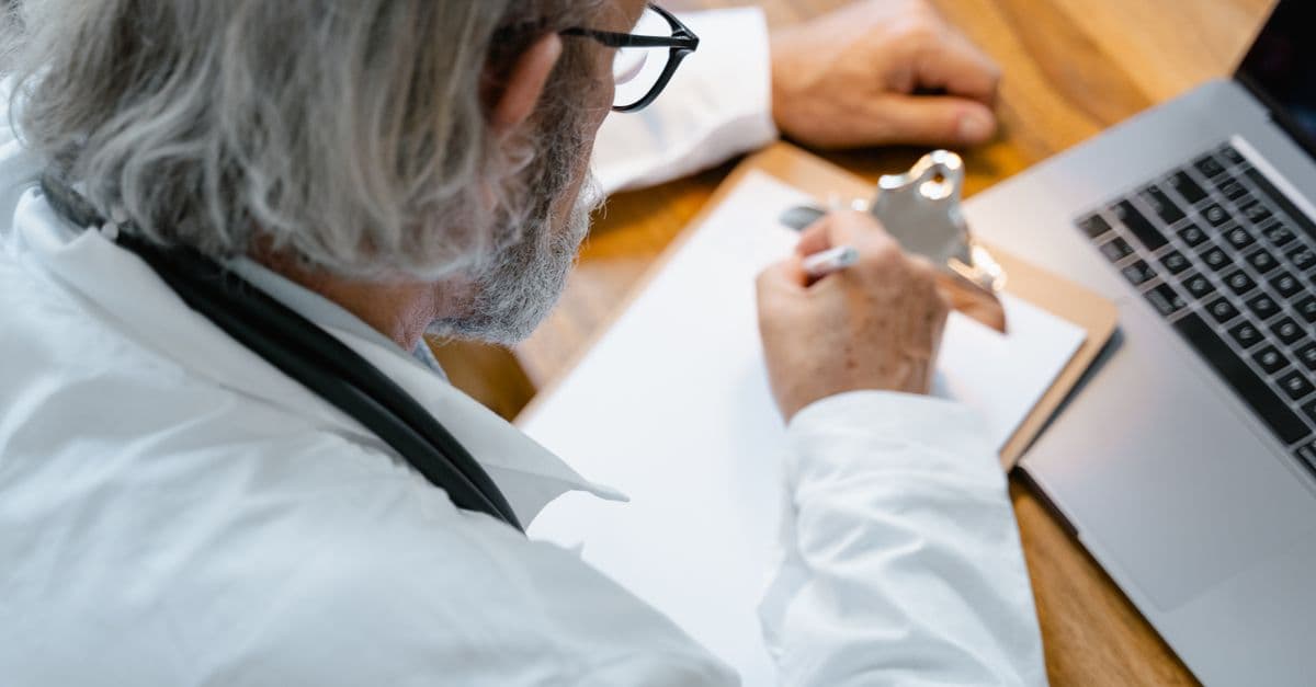 An elderly doctor in a white coat writing notes on a clipboard at a desk with a laptop.