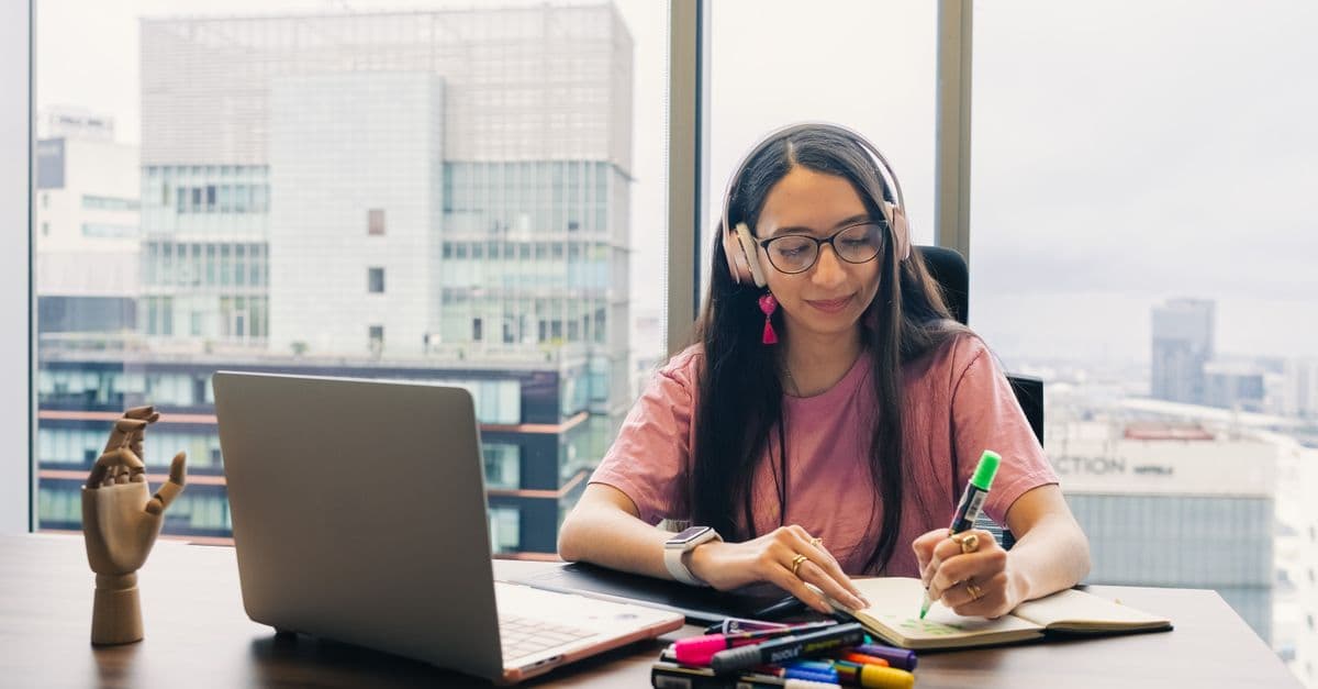 Young woman wearing headphones, writing in notebook at office desk with city view.