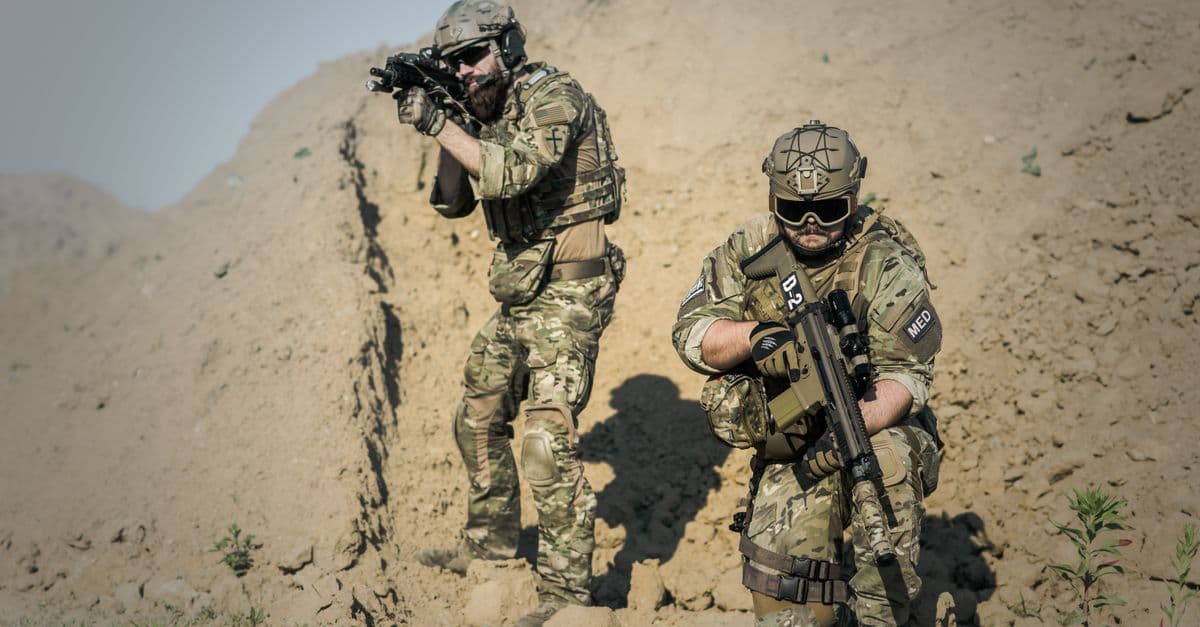 Two soldiers in camouflage gear equipped with weapons in a desert environment.