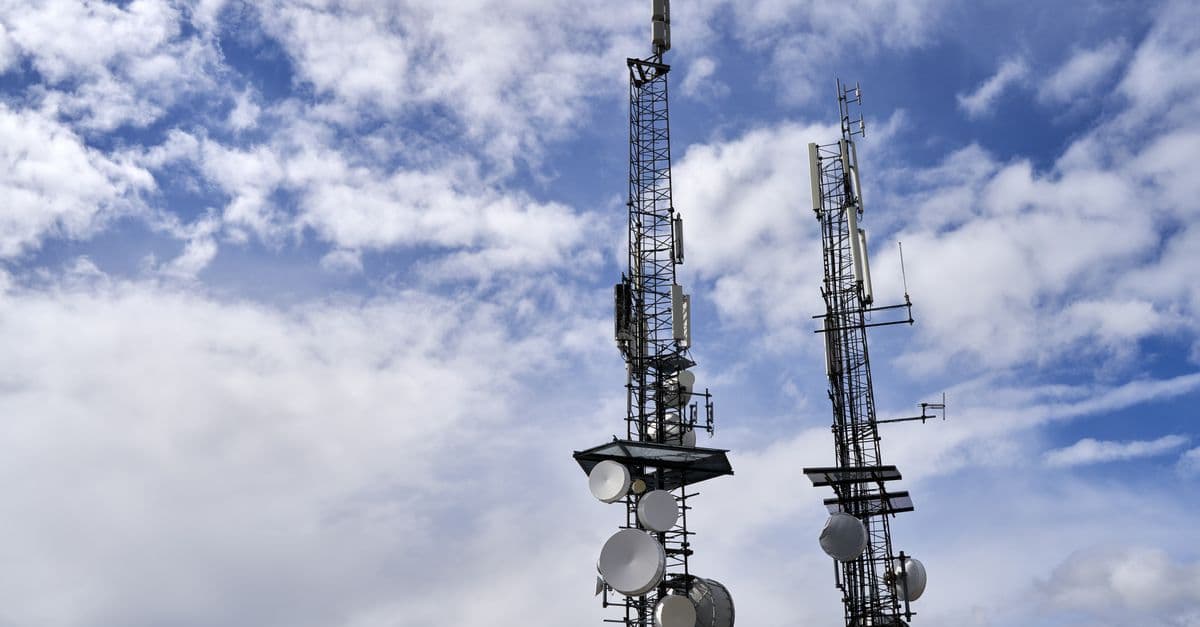 Telecommunication towers with antennas and dishes under a bright cloudy sky.