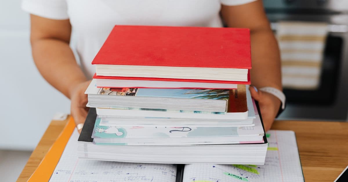 Close-up of a person holding a stack of colorful books over an open notebook.