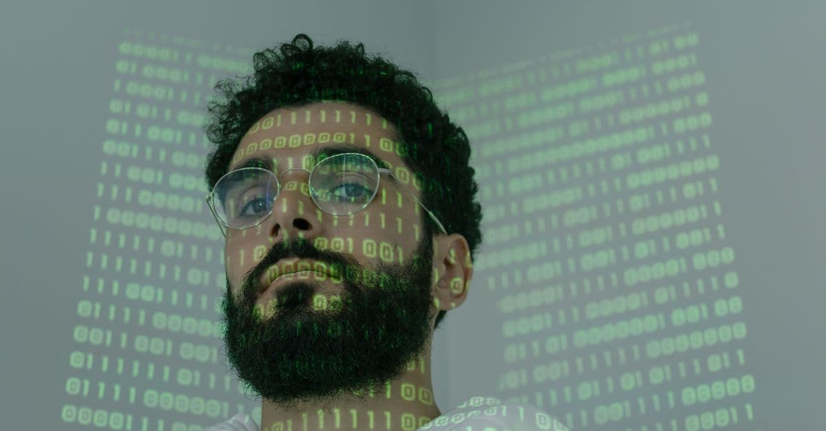 A man with a beard and eyeglasses looking at the camera, illuminated by binary code projection.