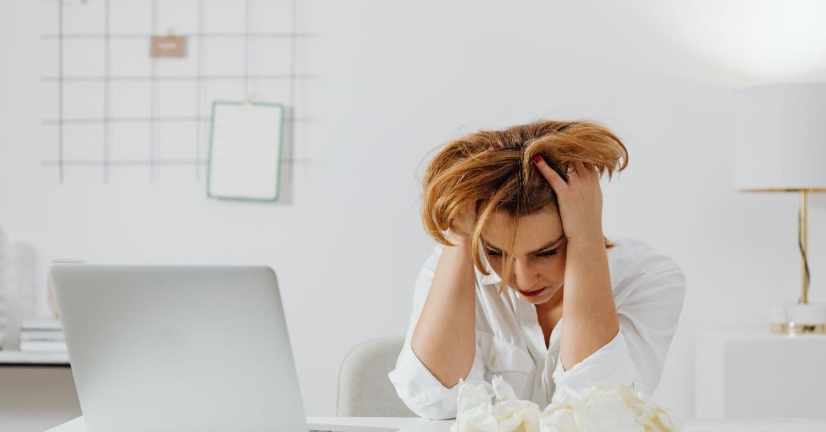 Frustrated woman in white shirt at desk with laptop, feeling overwhelmed.