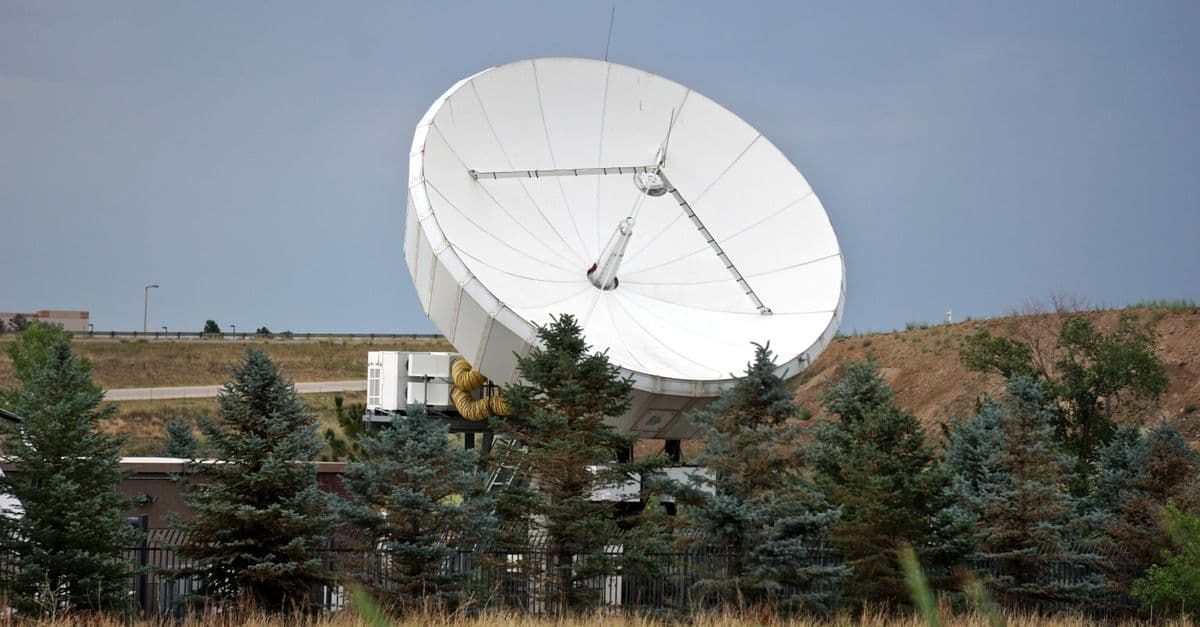 Wide shot of a satellite dish against a clear sky, surrounded by trees and grass.