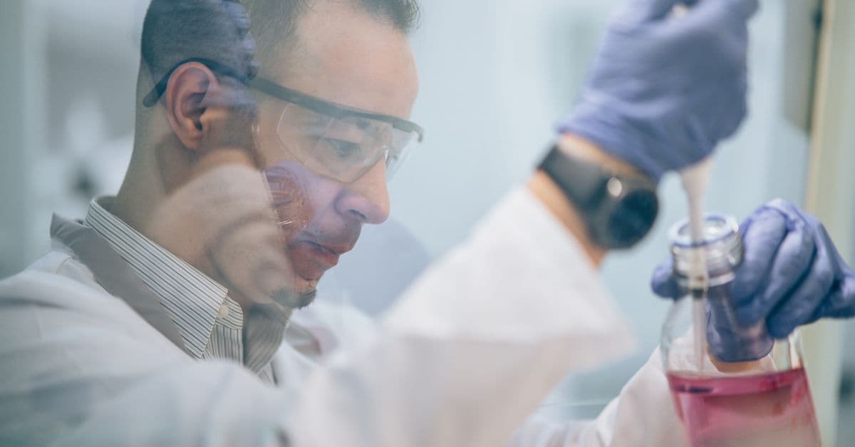 Scientist in lab coat and gloves transferring pink liquid using pipette.