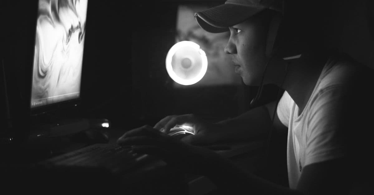 Side view of a gamer wearing a cap and headphones, illuminated by a computer screen in a dark room.