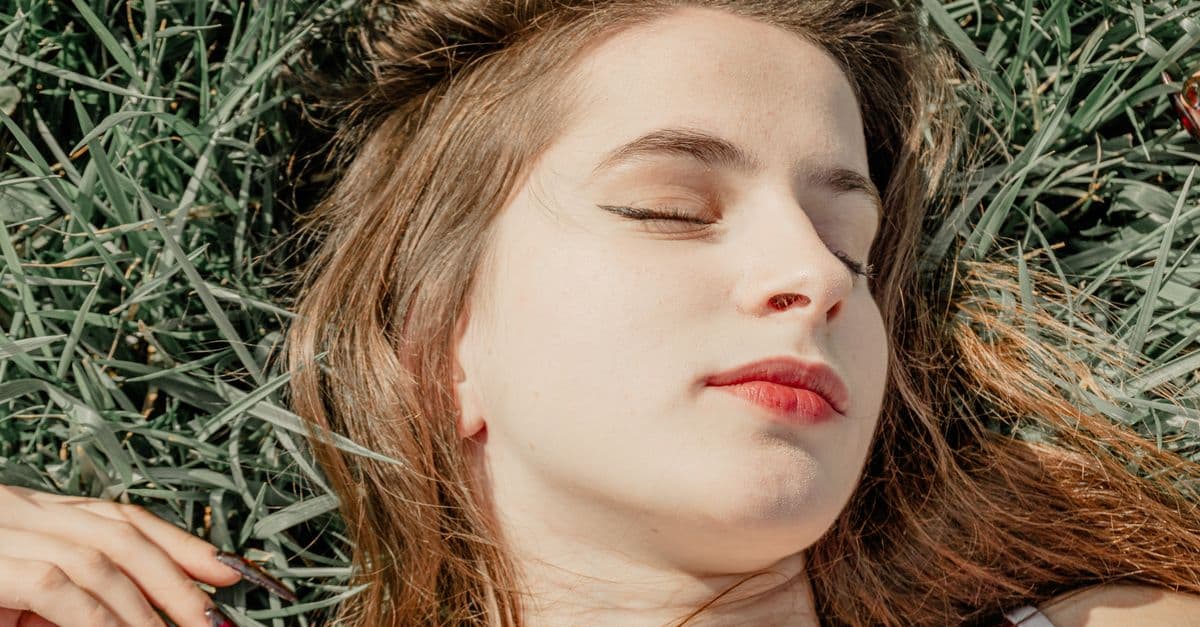 Portrait of a young woman with eyes closed lying on grass, enjoying sunlight.