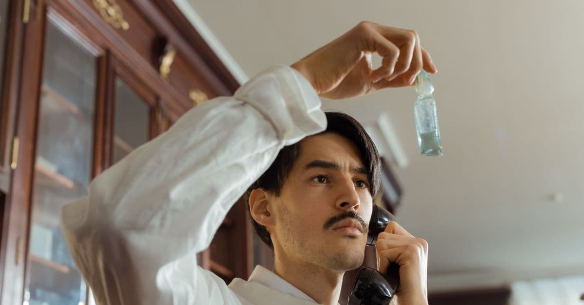 A man carefully examines a bottle sample while speaking on a vintage rotary phone indoors, symbolizing multitasking.