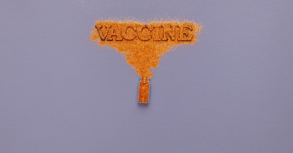 Orange glitter powder spreading into the word 'Vaccine' from a vial on a gray background.
