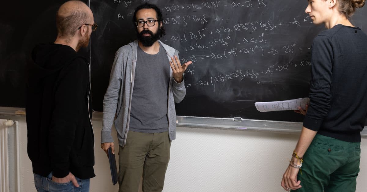 Three adults solving complex mathematical equations on a chalkboard indoors.