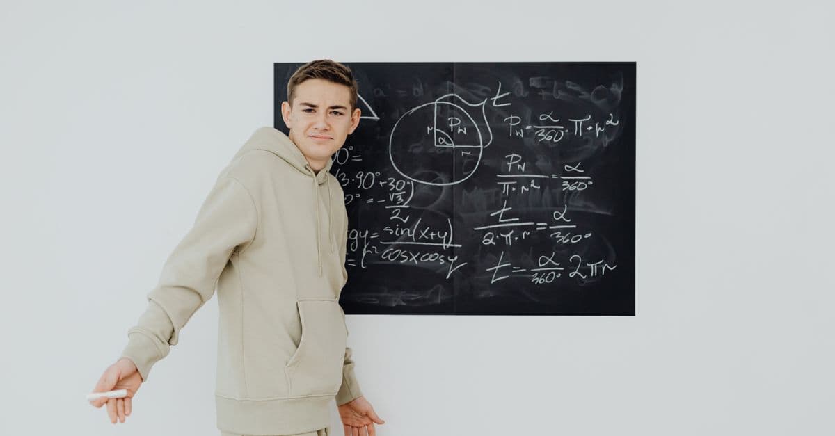 Teenager in casual clothes solving math equations on a blackboard indoors.