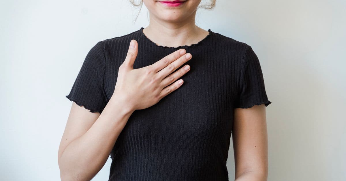 Asian woman in a black shirt expressing gratitude with a hand gesture against a neutral background.