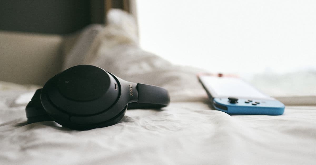 Close-up of a black headset and a gaming console resting on a soft, white bedspread in a calm, indoor setting.