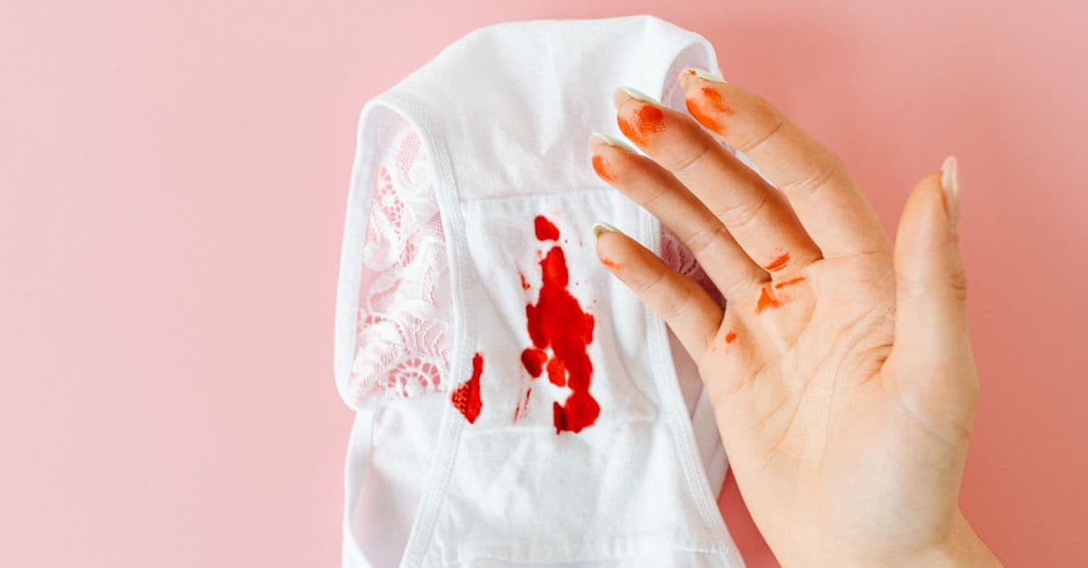 Close-up of a hand with blood stains next to bloodstained underwear on pink background.