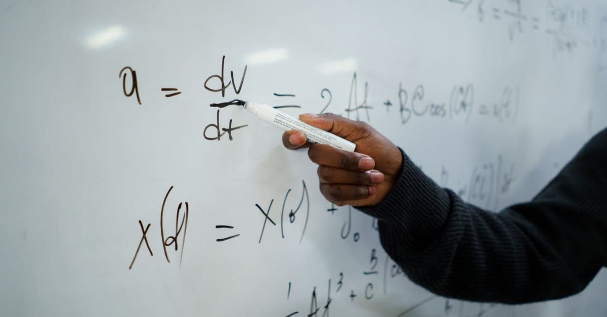 Close-up of hand writing equations on a whiteboard in a classroom setting.