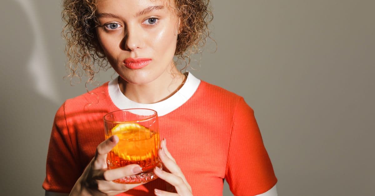 A woman in a red top holds a Negroni cocktail with an orange slice, in a well-lit setting.