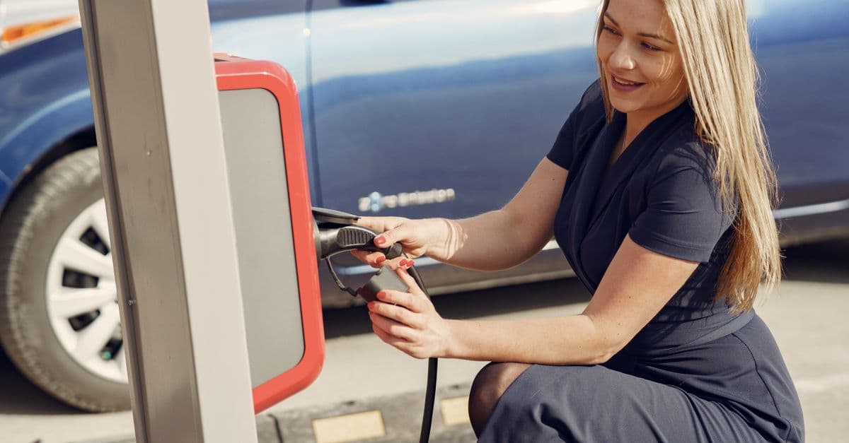 Side view of positive female in casual dress sitting near station on street charging electric vehicle for continuation trip