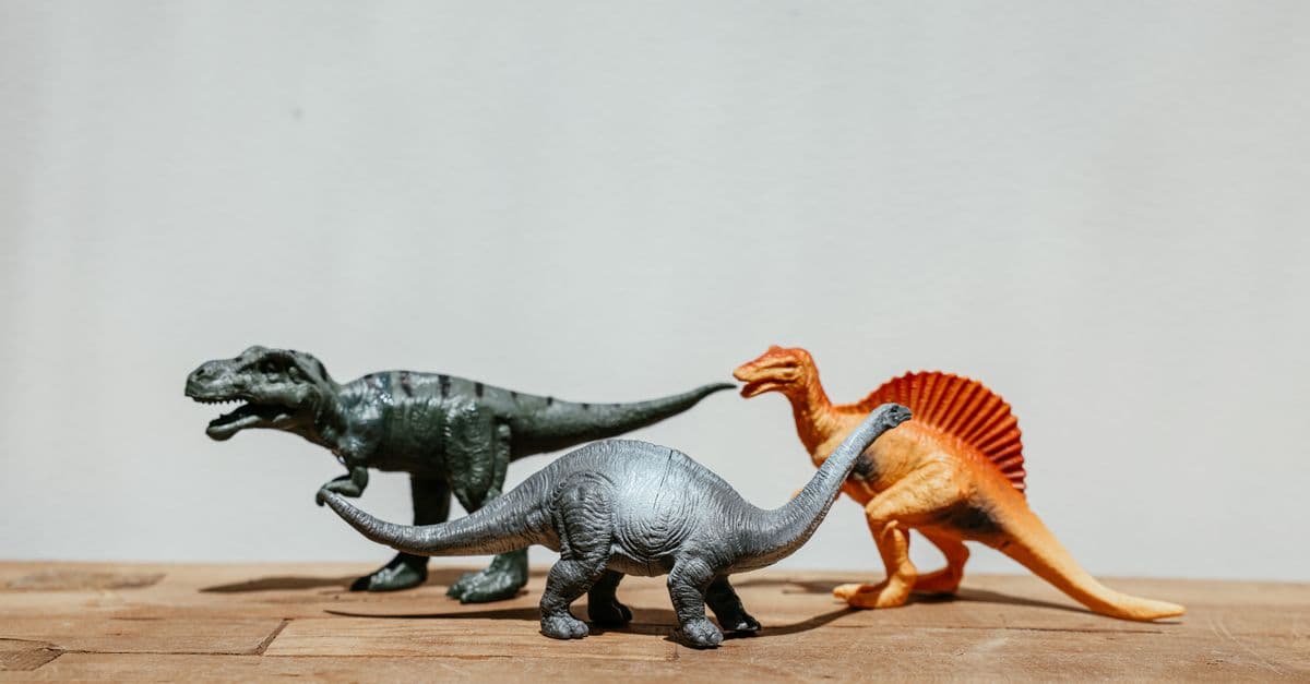 Three dinosaur toy figures displayed on a wooden board against a neutral background.