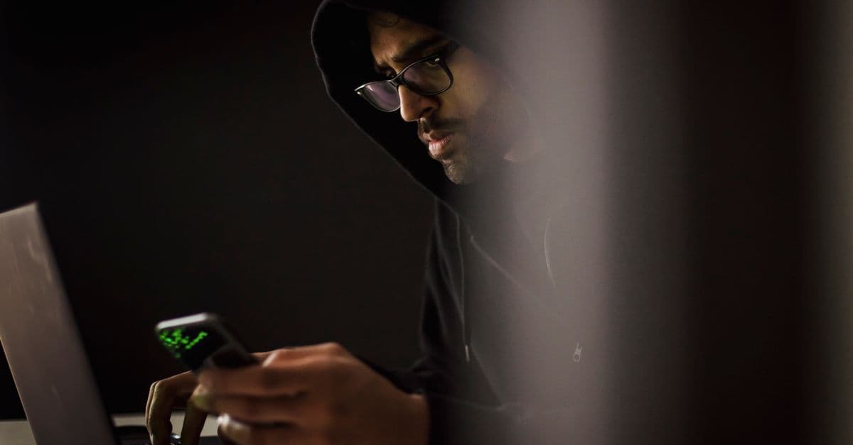 Crop bearded ethnic male computer geek in hoodie chatting on cellphone while typing on netbook in dark room