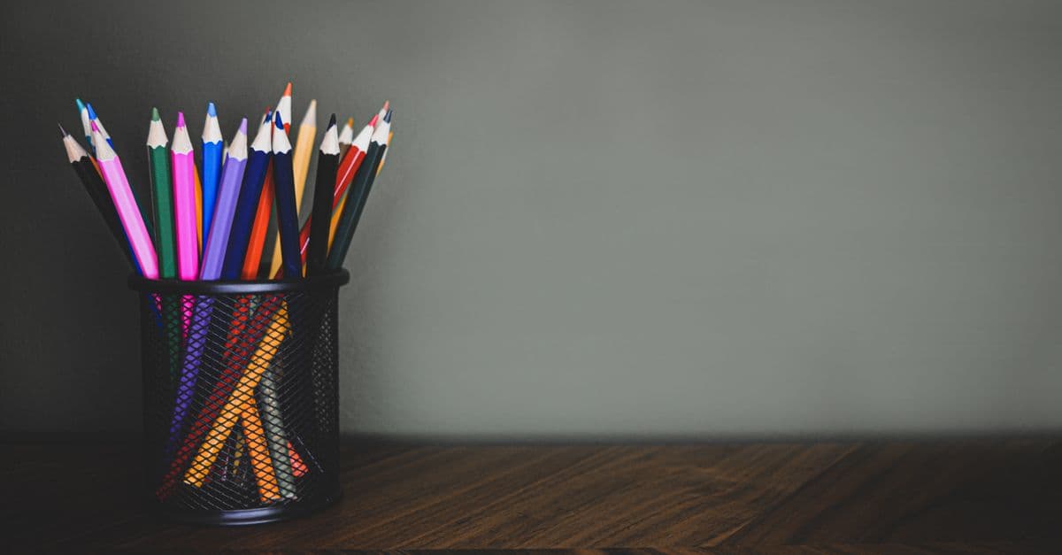 Vibrant color pencils in a black mesh holder placed on a dark wooden desk with ample copyspace.