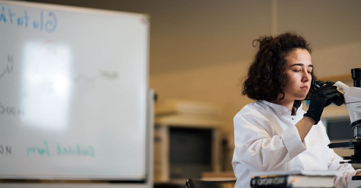 A female scientist in a lab coat examines a sample under a microscope in a laboratory setting.