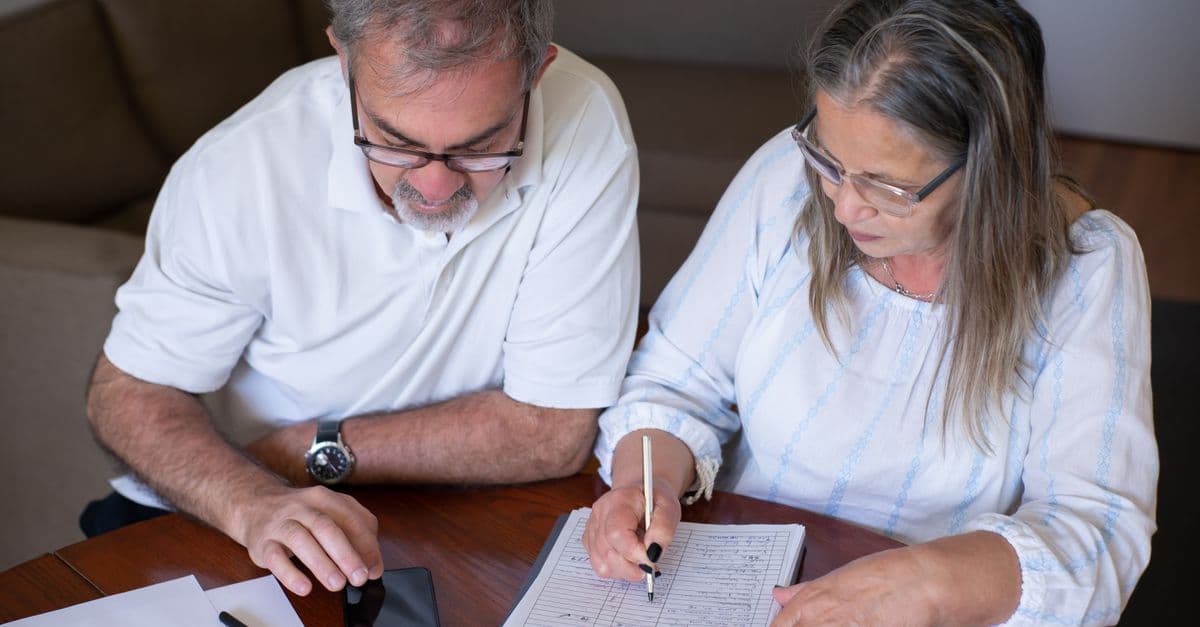 Elderly couple reviewing documents, using smartphone for online banking at home.