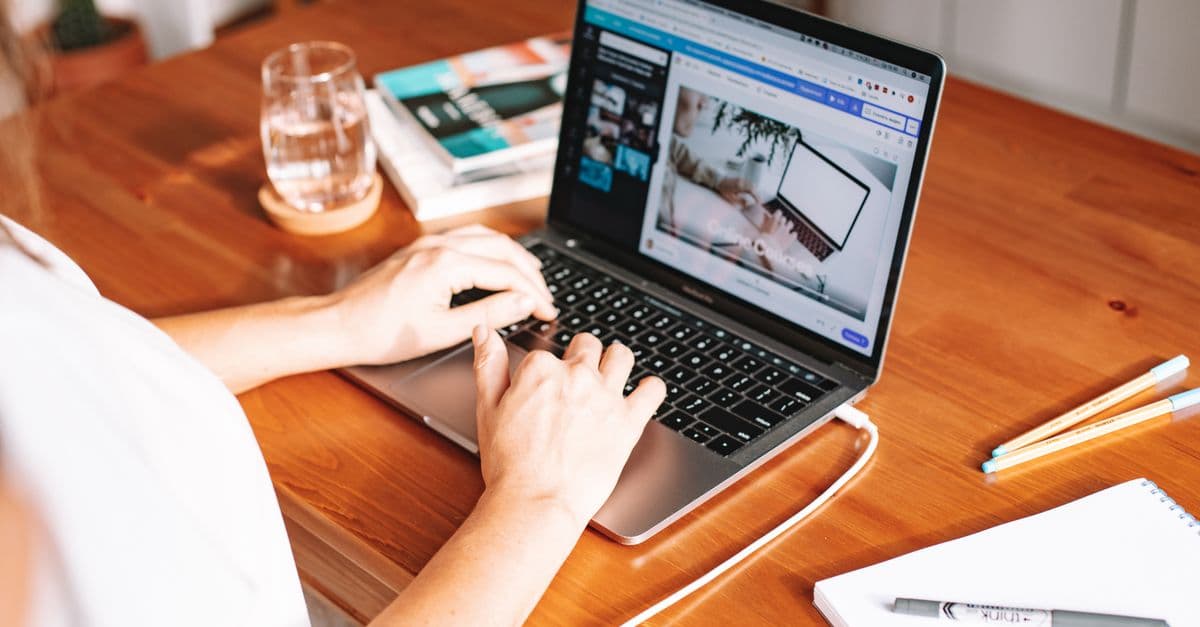 A person working remotely on a laptop at a wooden desk with stationery and a notepad.