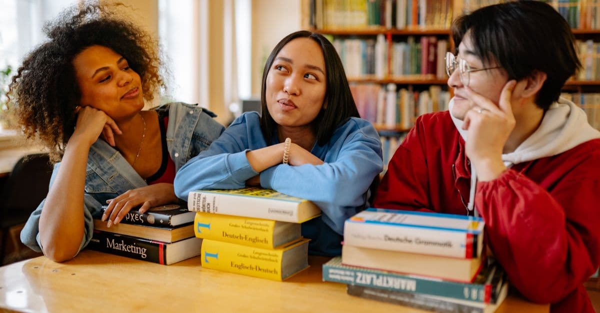 Three diverse students studying together in a university library with books around them.