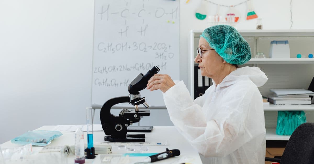 Senior scientist using a microscope in a laboratory setting wearing protective gear.
