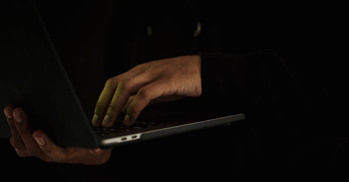 Crop anonymous male wearing black hoodie working on modern netbook while standing in dark studio
