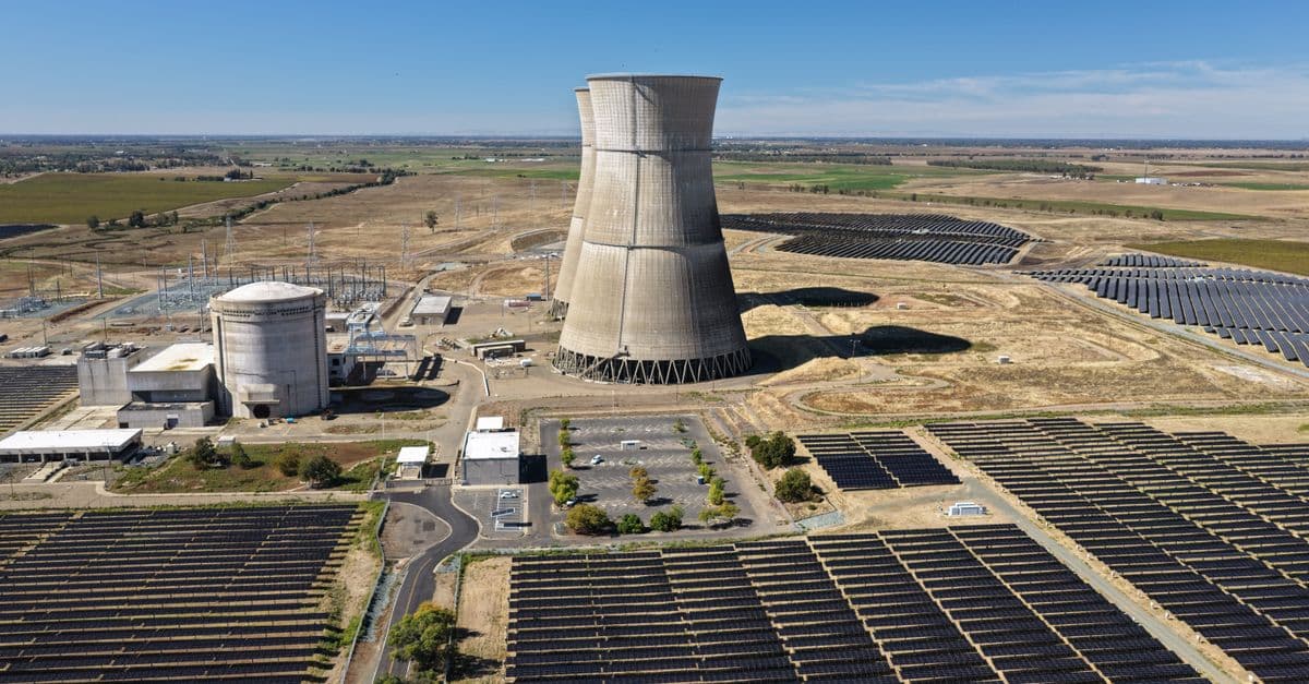 Aerial view of a nuclear cooling tower surrounded by solar panel fields in a rural landscape.
