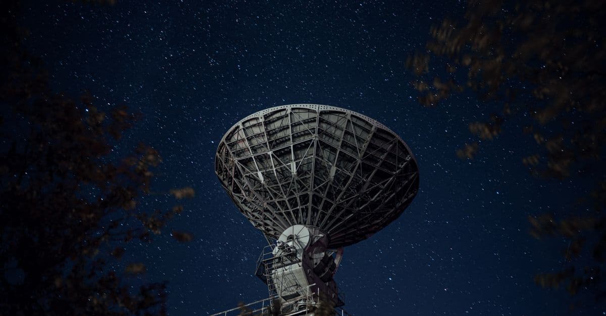 From below of blue starry sky over radio telescope and trees with leaves