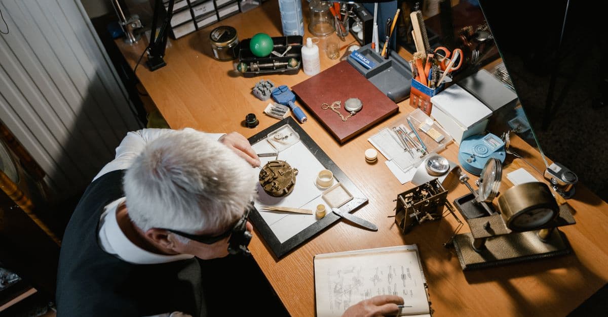 Elderly man focused on fixing a timepiece at a cluttered workbench with precision tools.