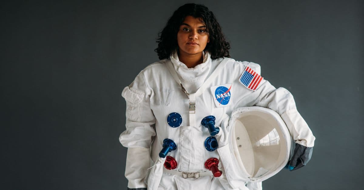 Woman dressed as astronaut holding helmet in NASA suit against grey background.
