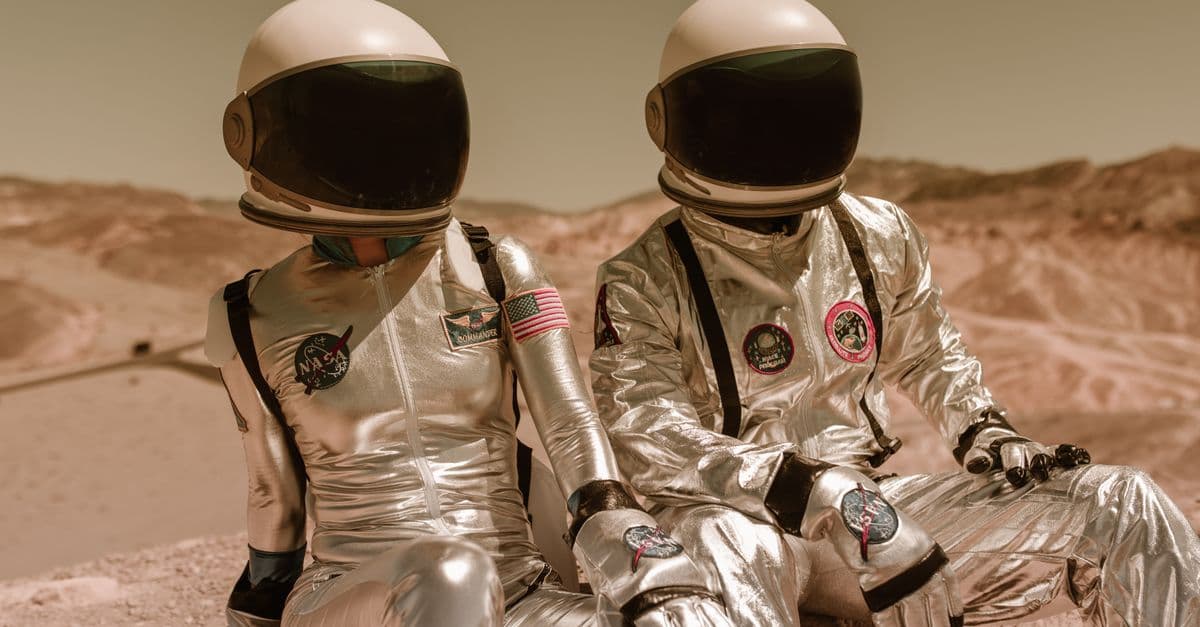 Two astronauts in silver suits and helmets sitting on a Mars-like desert landscape.