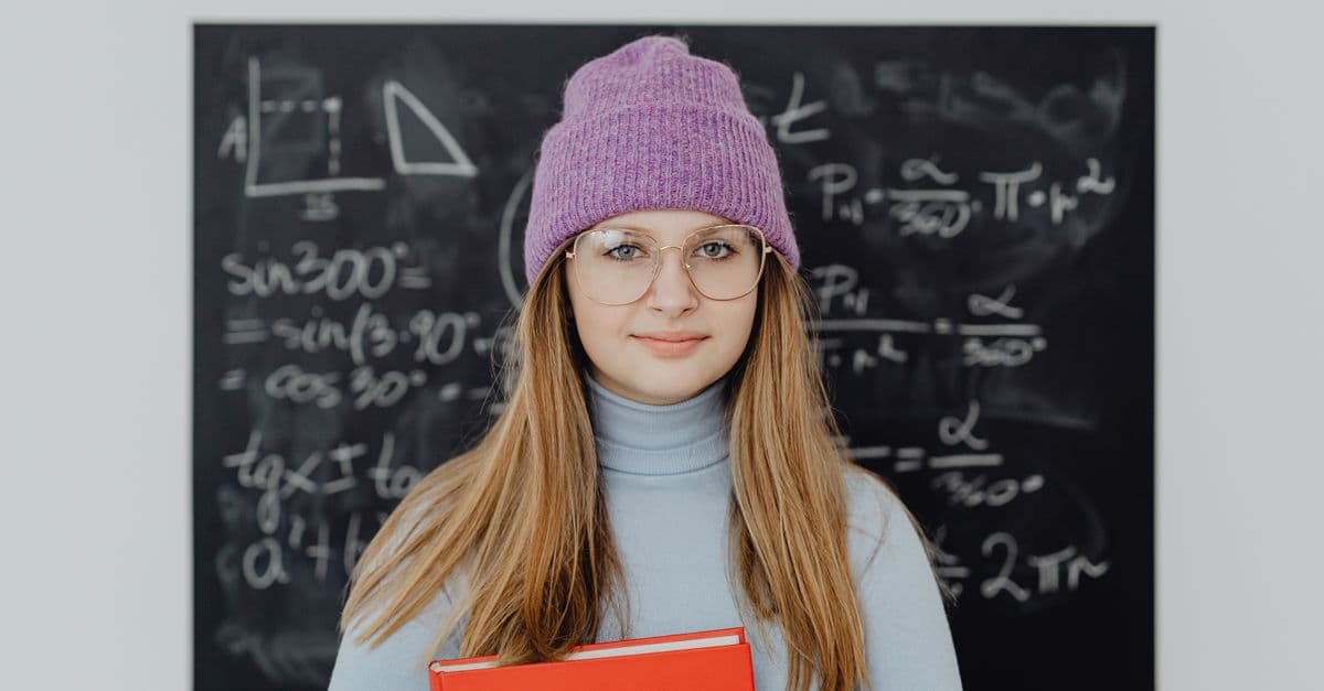 Teenage girl wearing glasses and a beanie, studying math indoors with a blackboard background.