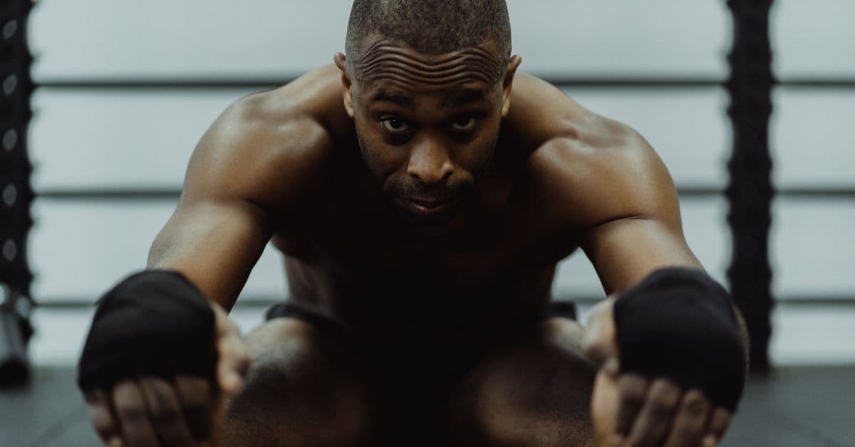 Topless African American man with hand wraps stretching in a gym environment.